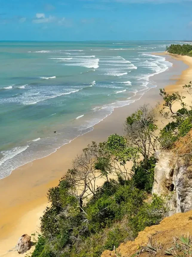 Uma vista panorâmica da costa em Arraial d'Ajuda mostra uma praia arenosa abraçada por ondas suaves de cor azul-turquesa. A costa é adornada com uma vegetação luxuriante e falésias com vista para a água, tudo sob um céu azul brilhante pontilhado por nuvens ocasionais. Ideal para casas de praia de alto padrão, afins às de Trancoso, projetadas por arquitetos renomados como Guilherme Torres.
