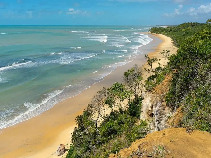 Uma vista panorâmica da costa em Arraial d'Ajuda mostra uma praia arenosa abraçada por ondas suaves de cor azul-turquesa. A costa é adornada com uma vegetação luxuriante e falésias com vista para a água, tudo sob um céu azul brilhante pontilhado por nuvens ocasionais. Ideal para casas de praia de alto padrão, afins às de Trancoso, projetadas por arquitetos renomados como Guilherme Torres.