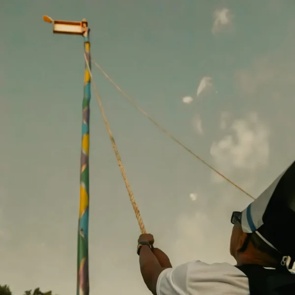 In Arraial d'Ajuda, beneath a cloudy sky, an individual in the foreground, donning a hat and glasses, is part of a group pulling on a rope tied to a tall pole with vibrant colors and topped by a small structure. This scene unfolds near the luxurious casas de praia de alto padrão of Trancoso.