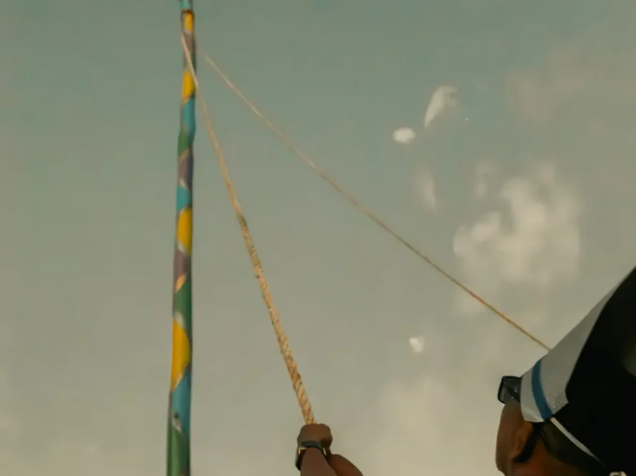 In Arraial d'Ajuda, beneath a cloudy sky, an individual in the foreground, donning a hat and glasses, is part of a group pulling on a rope tied to a tall pole with vibrant colors and topped by a small structure. This scene unfolds near the luxurious casas de praia de alto padrão of Trancoso.